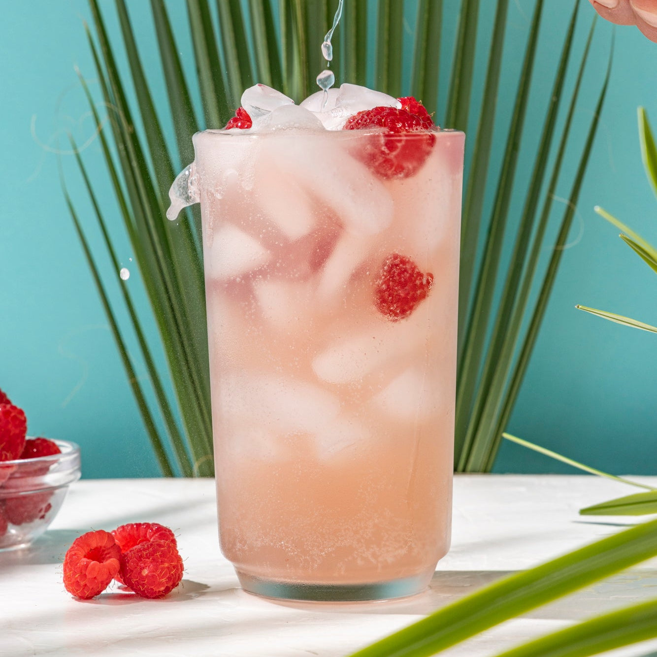 Person pouring a drink from a High Rise Raspberry THC Can into a glass with ice and raspberries, against a light blue background with palm leaves.