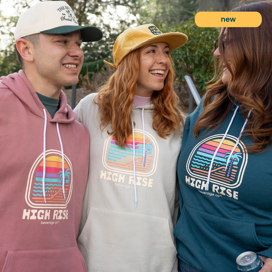 Three people wearing High Rise hoodies and caps outdoors. The male on the left has the mauve hoodie on, the female in the center has the cream hoodie on and the woman on the right has the navy hoodie on.