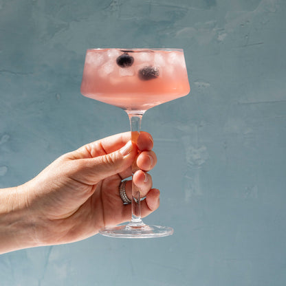 Hand holding a blueberry High Rise Seltzer with blueberries in a glass against a blue background