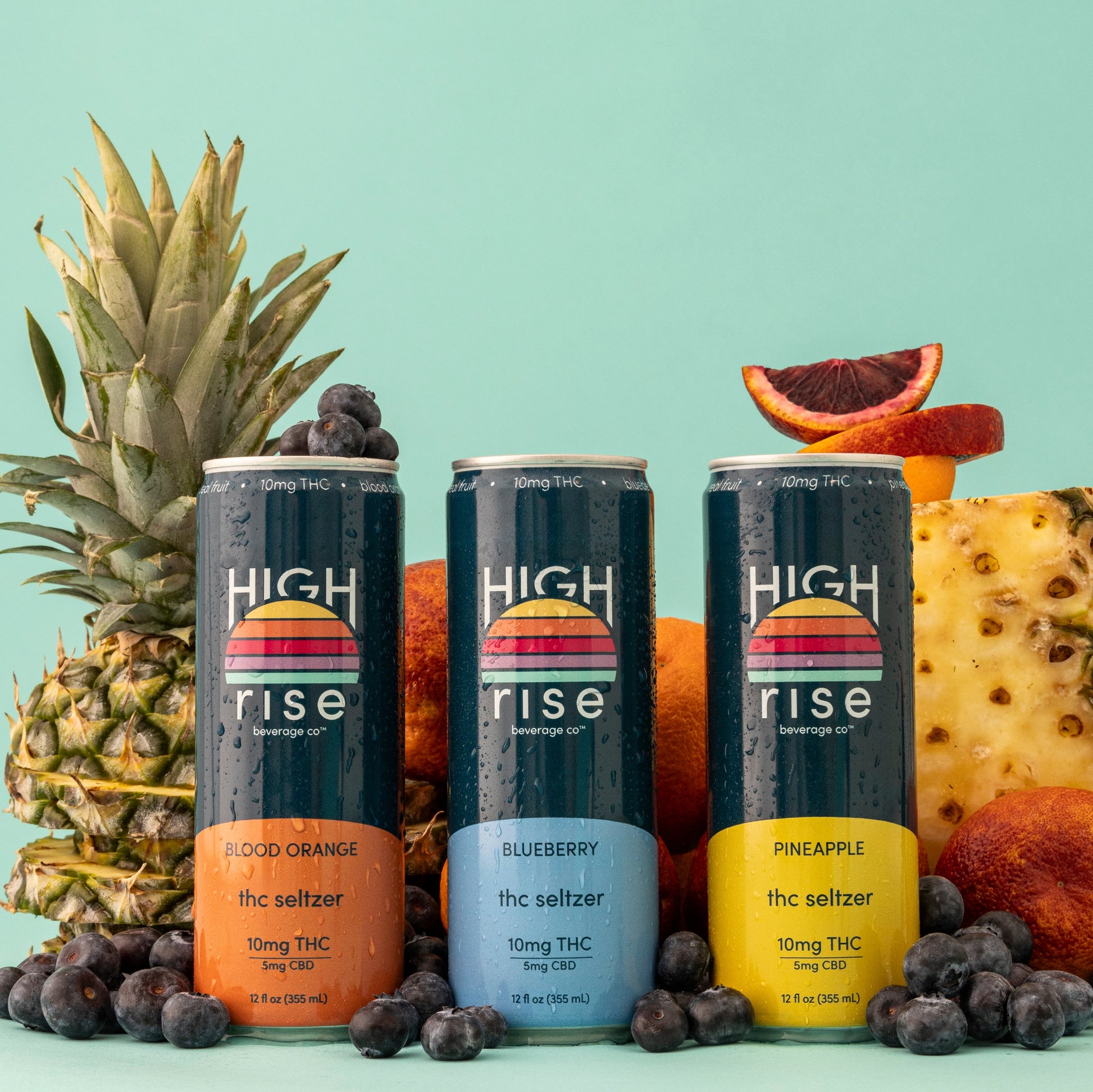 Three cans of 'High Rise' THC-infused drinks surrounded by pineapples, oranges, and blueberries on a turquoise background.