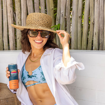 Woman in a bikini and straw hat holding a can of High Rise Blood Orange 10mg THC Seltzter, sitting on a wooden bench.