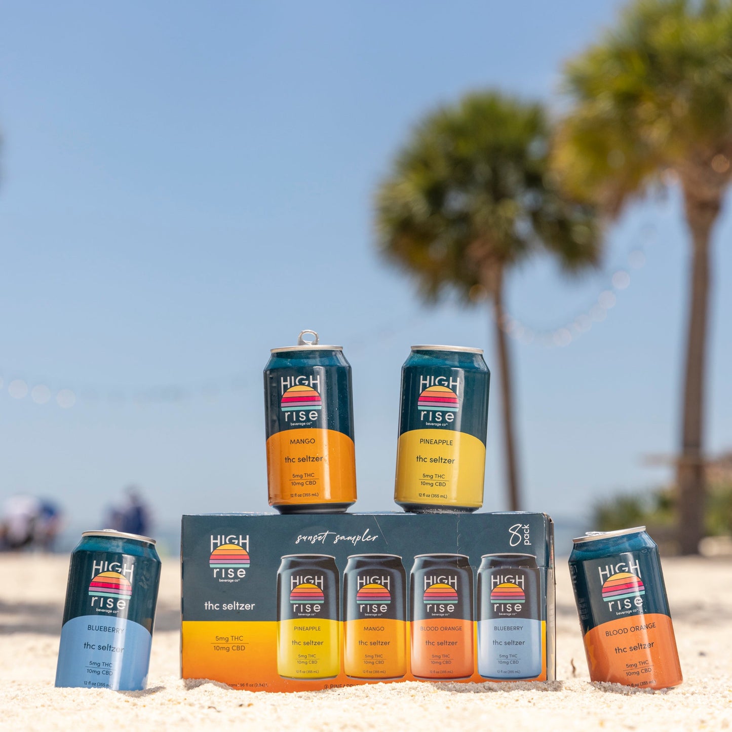 Cans of High Rise THC 5mg Seltzer on a sandy beach with palm trees in the background. 